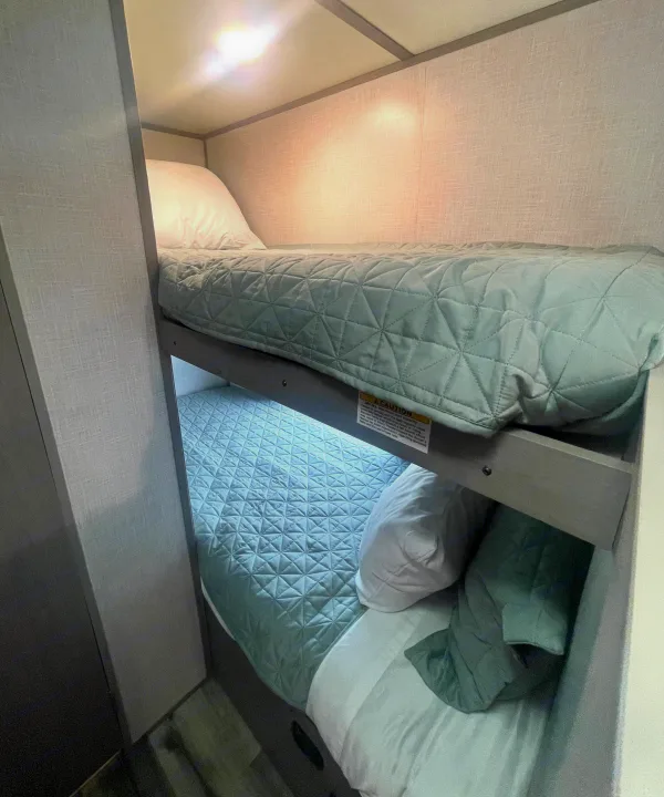 Comfy RV rental bunk beds feature light green quilted blankets and white pillows, perfect for travel adventures.. Dutchmen Coleman Lantern LT 2023
