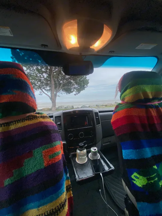 Inside an RV rental: two travelers cozy in colorful ponchos, enjoying a serene beach view. Lights are on overhead.. Airstream Interstate 2013