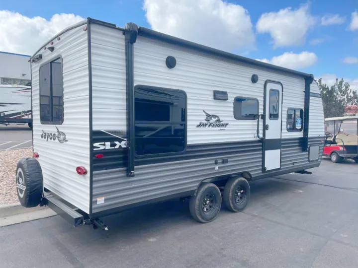 This spacious white and grey Jay Flight travel trailer, ideal for an RV rental, features an awning, windows, and a rear spare tire for your next outdoor adventure.. Jayco Jay Flight SLX 2024