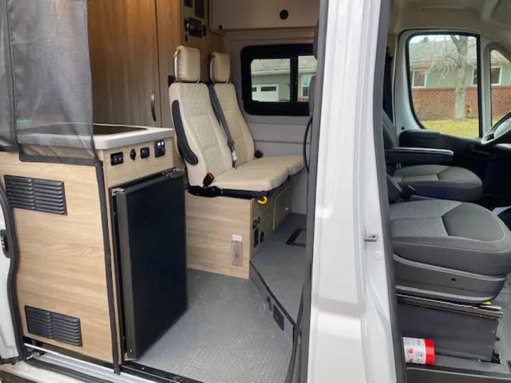 RV rental interior: A compact kitchen with a fridge is on the left, beside two beige passenger seats. Part of the front cab is visible.. Winnebago Solis 2021