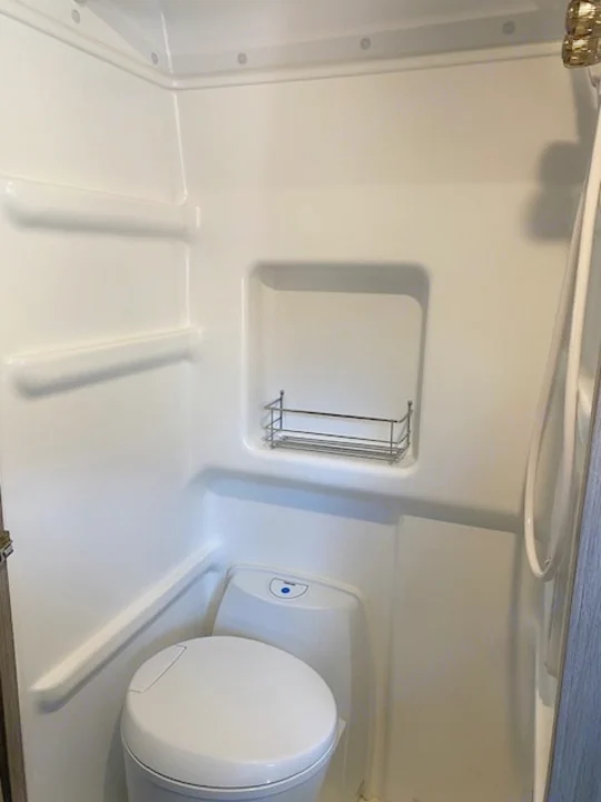 Compact, all-white RV rental wet bath featuring a toilet, integrated shower, and built-in shelving.. Winnebago Solis 2021