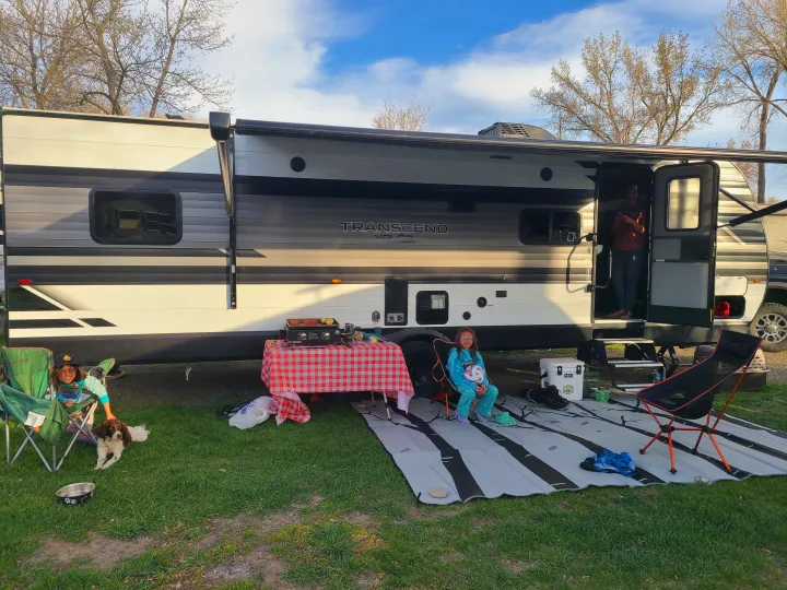 Relaxing RV rental camping. Family, kids, & dog enjoy outdoor cooking setup by their travel trailer under the awning.. Grand Design Transcend Xplor 2022
