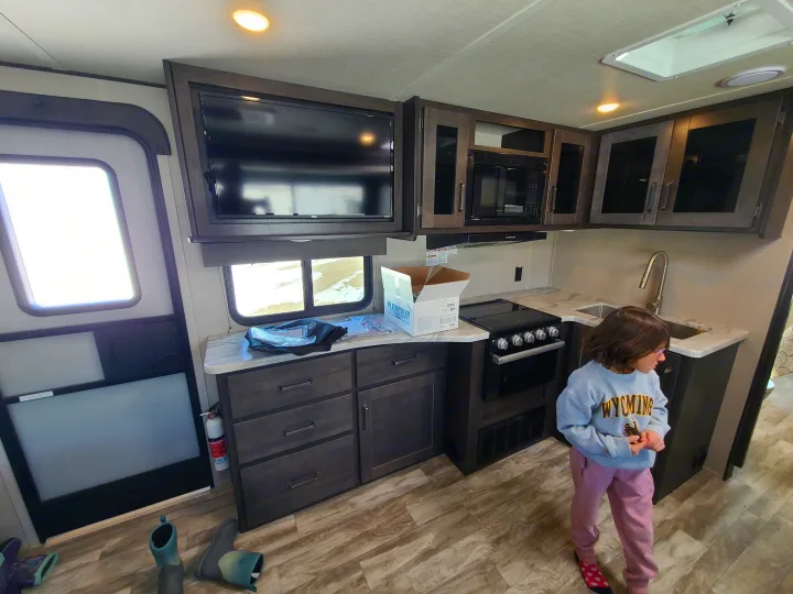 Inside a modern **RV rental**, a child explores the kitchen with stove, microwave, and TV setup.. Grand Design Transcend Xplor 2022