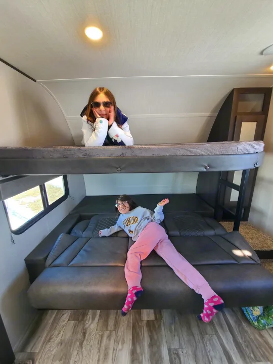 Inside an RV rental, two girls pose. One on the upper bunk with shades, the other on the lower bed in a Wyoming shirt.. Grand Design Transcend Xplor 2022