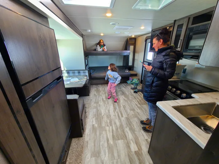 Family in an RV rental: An adult looks at her phone while children play, showing the kitchen, fridge, and bunk beds.. Grand Design Transcend Xplor 2022