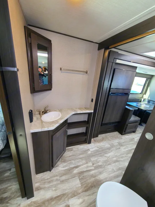 RV rental interior: Bathroom vanity with sink, medicine cabinet, and kitchen/dining area with a fridge. Features dark wood and light flooring.. Grand Design Transcend Xplor 2022