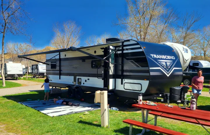 Experience an RV rental! A Transcend Xplor travel trailer set up in a sunny campground with a family relaxing.. Grand Design Transcend Xplor 2022