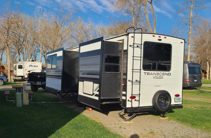 A white Grand Design Transcend XPLOR RV rental with extended slide-outs is parked in a sunny campground, with visible utility hookups.. Grand Design Transcend Xplor 2022