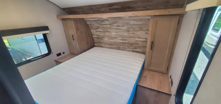 Inside an RV rental bedroom: bed with white mattress, wood headboard, cabinets, and a window for natural light.. Gulf Stream Trailmaster 2023