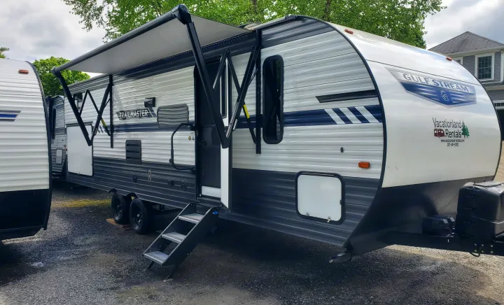 A grey & white Gulf Stream travel trailer RV rental, with awning extended & steps down, parked on gravel. Another RV is on the left.. Gulf Stream Trailmaster 2023