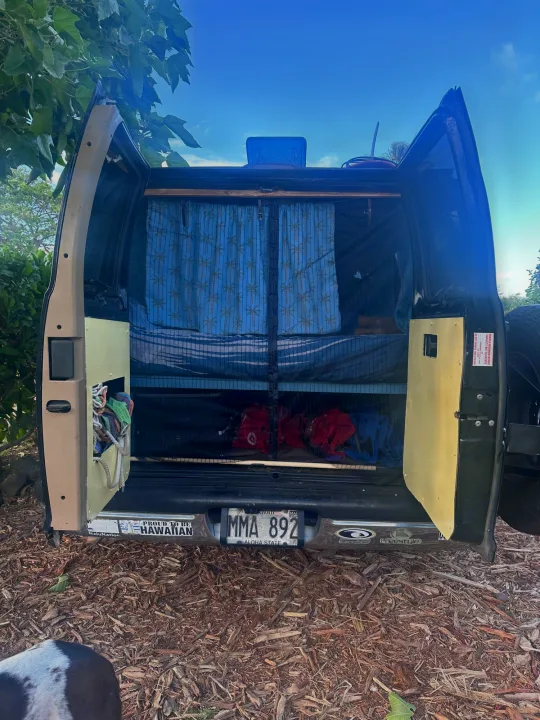 Open rear doors of a DIY RV rental van conversion, revealing a bed, storage, and tropical curtains. Hawaiian details visible.. Dodge 1500 1999