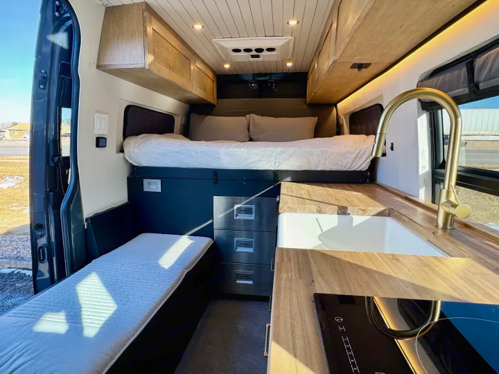 Inside a modern RV rental: bed, wooden cabinets, kitchen with sink, cooktop, and fridge.. Mercedes-Benz Sprinter 2021