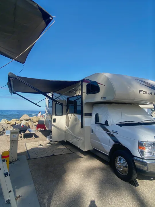 Serene RV rental campsite at the beach. A Four Winds RV is set up with awnings, overlooking the ocean with a person relaxing.. Thor Four Winds 2020