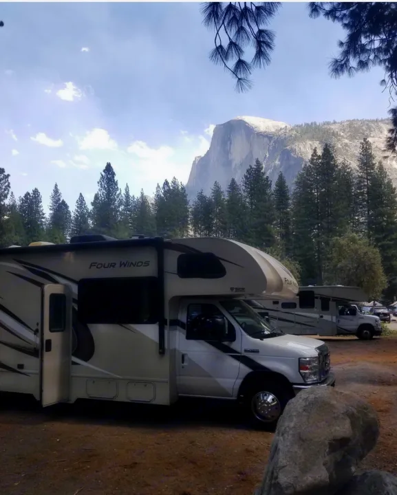 A white Four Winds RV rental is parked at a Yosemite campground, with Half Dome and pine trees in the background.. Thor Four Winds 2020