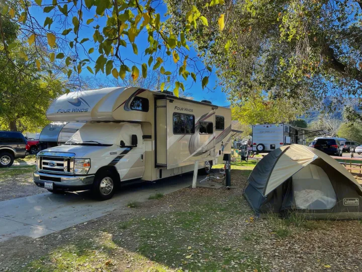 This sunny image shows an RV rental and a small tent set up at a busy campground, offering outdoor adventure and camping fun.. Thor Four Winds 2020
