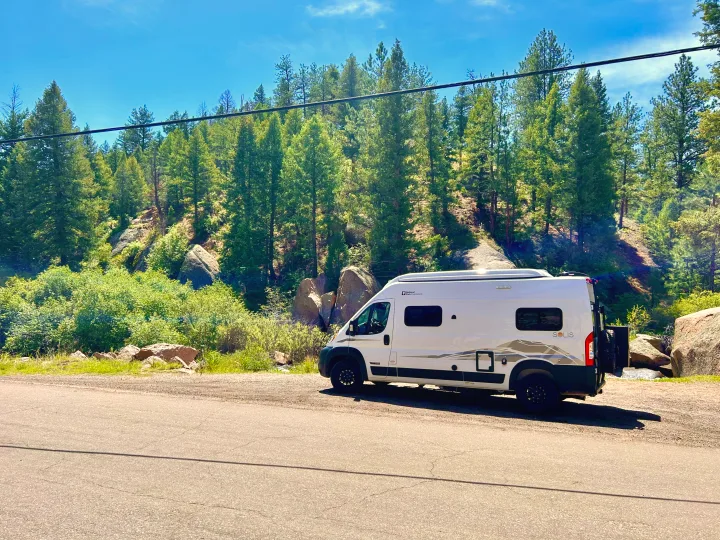 White Winnebago Solis RV rental parked on a mountain road. Sunny forest, rocks, and blue sky create a scenic backdrop.. Winnebago Solis 2023