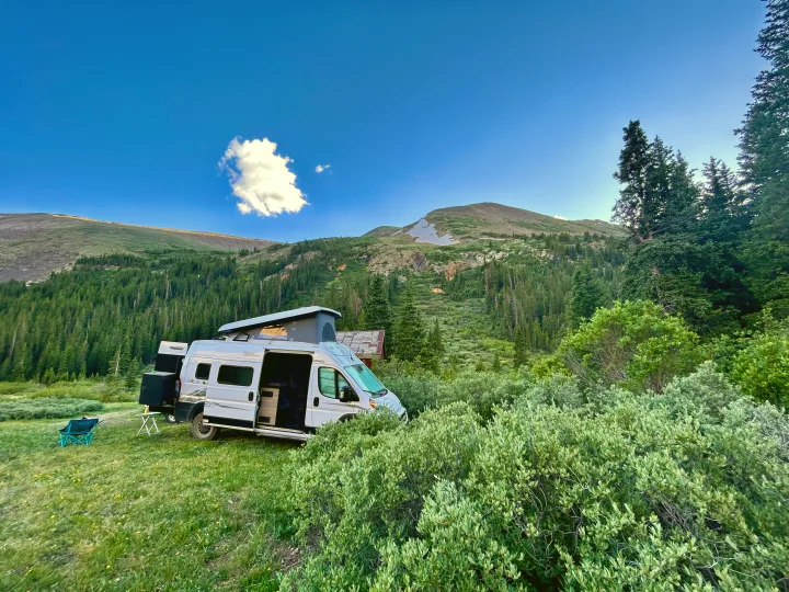 RV rental camper van with pop-top raised, nestled in a mountain forest clearing under a blue sky.. Winnebago Solis 2023