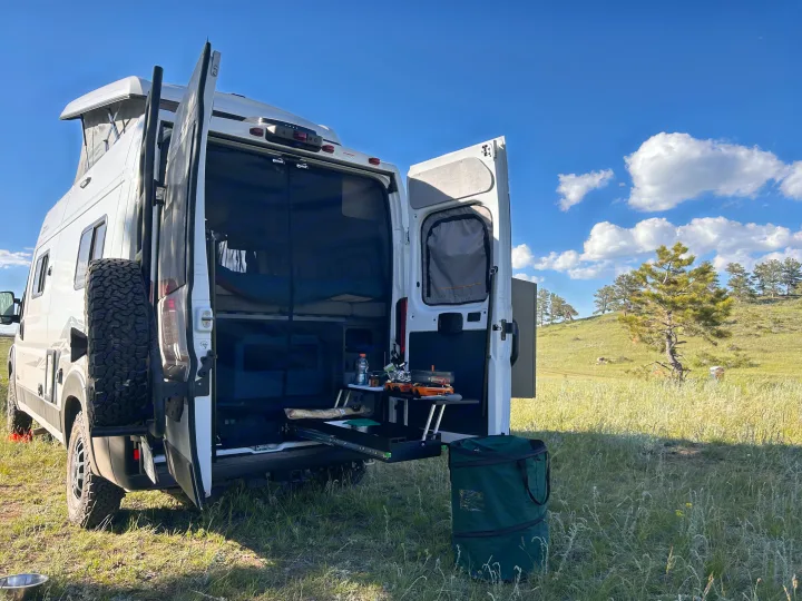 An RV rental with open doors, revealing an organized van life setup for cooking & camping in a scenic grassy field.. Winnebago Solis 2023