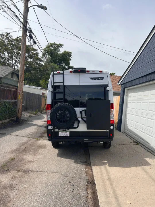 Rear view of a white Winnebago rv rental camper van with a ladder, spare tire, and black storage box, parked in an alley.. Winnebago Solis 2023