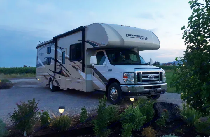 A Freedom Elite Class C rv rental, a beige motorhome, parked on gravel at dusk, with lit solar lights and green fields.. Thor Freedom Elite 2017