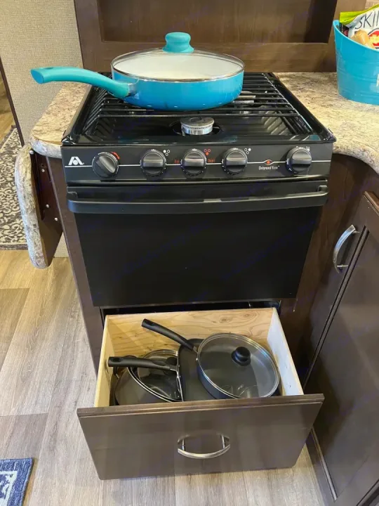 RV rental kitchen: Black gas stove with blue pot; drawer below open, showing stored black pots and pans.. Thor Freedom Elite 2017