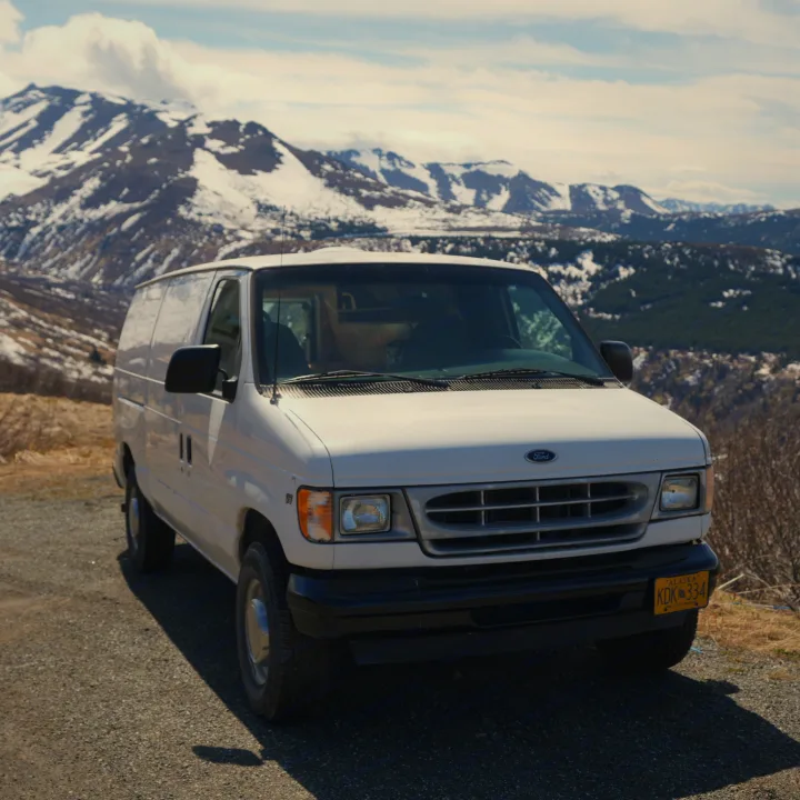 A white cargo van is parked on a gravel mountain road with snow-capped peaks in the background. Ideal for an Alaska RV rental adventure.. Ford E250 Camper Van 2001