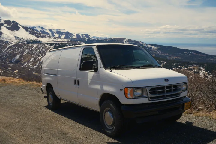 White Ford van parked on a scenic mountain road overlooking snowy Alaskan peaks and a bay, ideal for an RV rental adventure.. Ford E250 Camper Van 2001