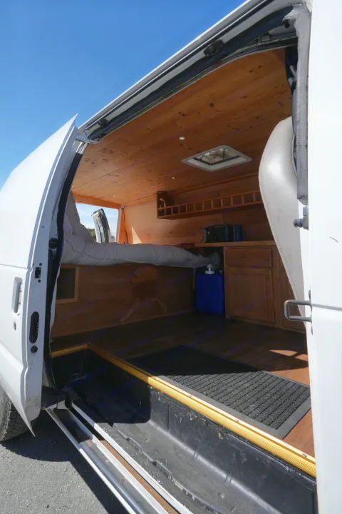 View inside an RV rental camper van: comfortable bed, wood-paneled walls/ceiling, kitchen area, and open side door.. Ford E250 Camper Van 2001