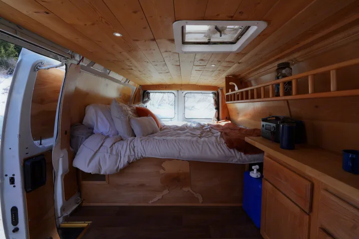 Cozy RV rental interior with wooden walls, comfy bed, and snowy landscape visible through the open van doors.. Ford E250 Camper Van 2001