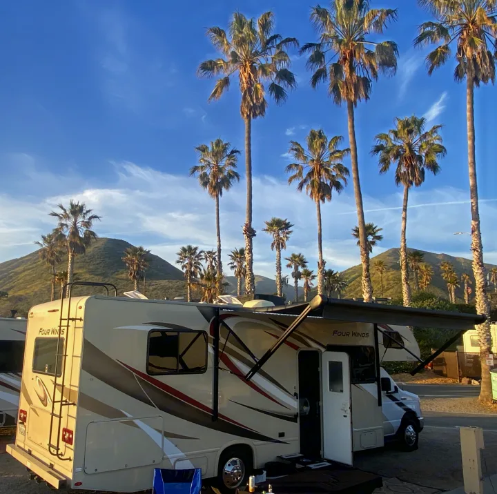 Spacious RV rental with an extended awning, parked among tall palm trees and hills under a blue sky, ideal for a getaway.. Thor Four Winds 2020