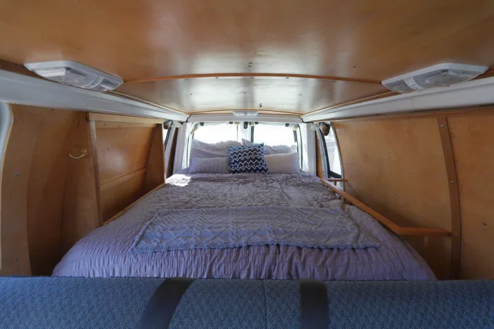 This cozy RV rental offers a bed inside a wood-paneled van, featuring ceiling lights and natural light from rear windows.. Chevrolet Other 2006