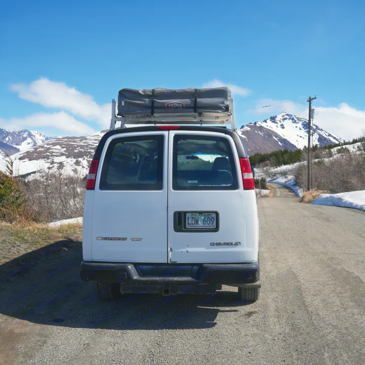 White cargo van RV rental with rooftop tent on a scenic gravel road in snowy Alaskan mountains under blue sky.. Chevrolet Other 2006