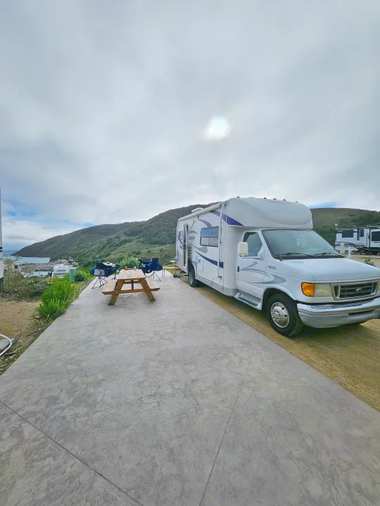 Enjoying the view from an RV rental campsite: a white motorhome parked next to a picnic table overlooks a bay with boats and green hills.. Coachmen Concord 2004