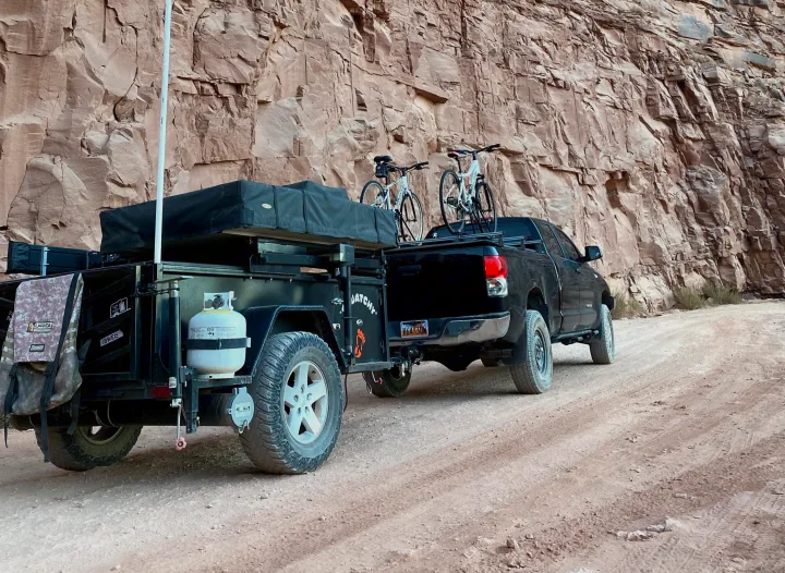 This RV rental setup is adventure-ready! A truck pulls a camping trailer with bikes on a dirt road by a towering canyon wall.. Insane Custom Trailers "Squatchy" 2022