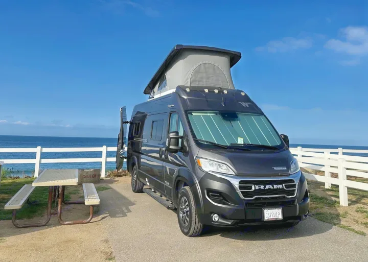 Dark grey RV rental camper van with pop-top, parked oceanfront with a picnic table. Scenic view for travel.. Winnebago Solis 2023