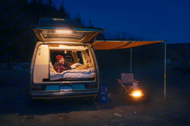 Relaxing RV rental night: Person inside a warm, lit camper van. Campfire glows outside near an extended awning.. Volkswagen T3 Westfalia 1985