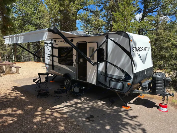 White Starcraft travel trailer rv rental set up at a shady campsite with an extended awning, chairs, and fire pit. Ideal for camping.. Starcraft Launch Outfitter Extreme 2018