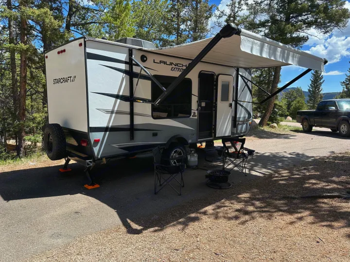 A modern Starcraft RV rental is parked at a serene campsite, with its awning extended, chairs, and fire pit ready for relaxation.. Starcraft Launch Outfitter Extreme 2018