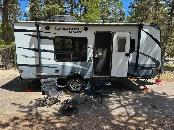 Enjoying the outdoors with an RV rental! A white travel trailer is set up with chairs and a fire pit on a shaded campsite.. Starcraft Launch Outfitter Extreme 2018