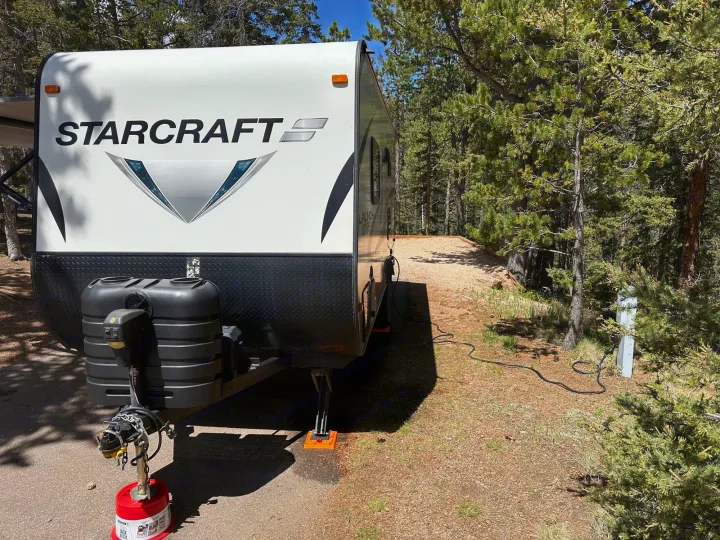 STARCRAFT RV rental parked among pine trees at a sunny campsite. Utility hookups visible, ready for travel.. Starcraft Launch Outfitter Extreme 2018