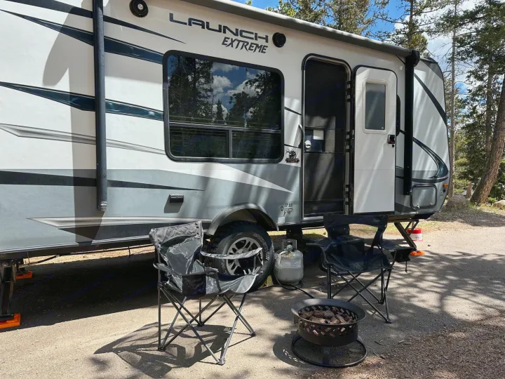 White and gray LAUNCH EXTREME RV rental parked in a wooded campsite with two chairs and a fire pit ready for relaxation.. Starcraft Launch Outfitter Extreme 2018