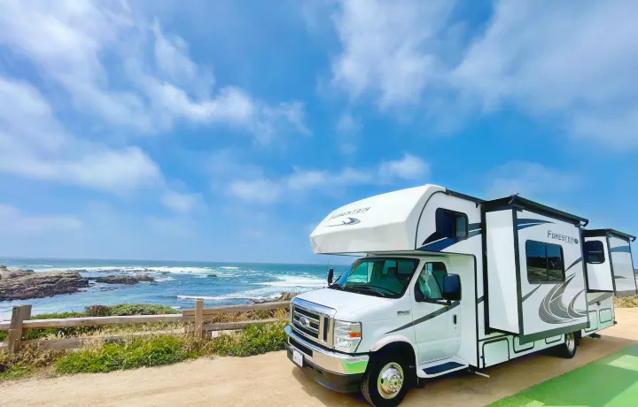 White RV rental parked at a scenic ocean overlook, waves crashing on rocks under blue sky. A dog peeks from the window.. Forest River Forester 2021