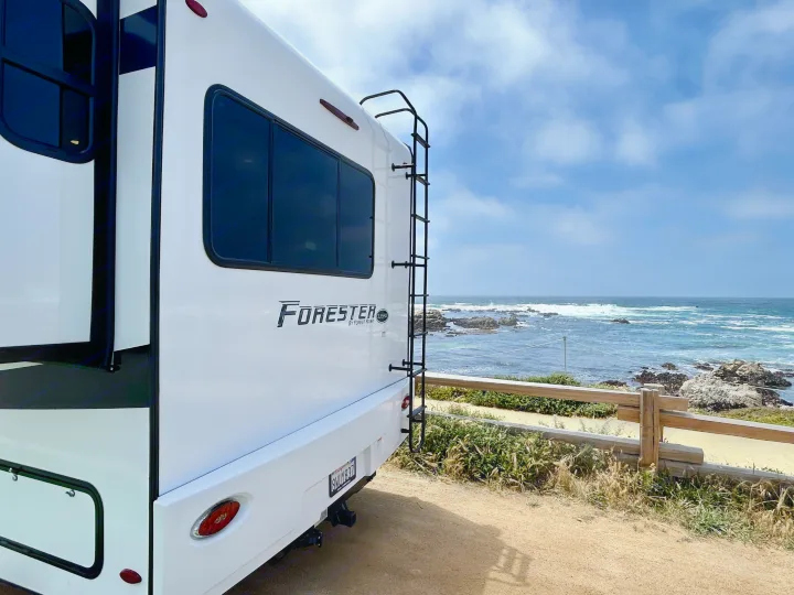 Rear of a white RV rental parked on a dirt path, offering a stunning view of the blue ocean and rocky coastline under a cloudy sky.. Forest River Forester 2021