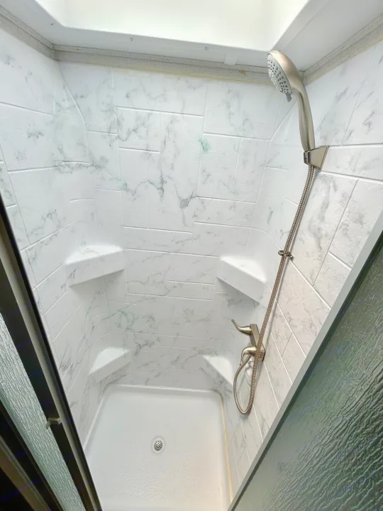 Clean and compact RV rental shower, featuring faux marble walls, a handheld showerhead, and built-in shelves.. Forest River Forester 2021