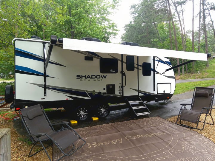 This Shadow Cruiser RV rental is set up at a green campsite with an extended awning, chairs, and a "Happy Campers" mat.. Cruiser RV Shadow Cruiser 2023