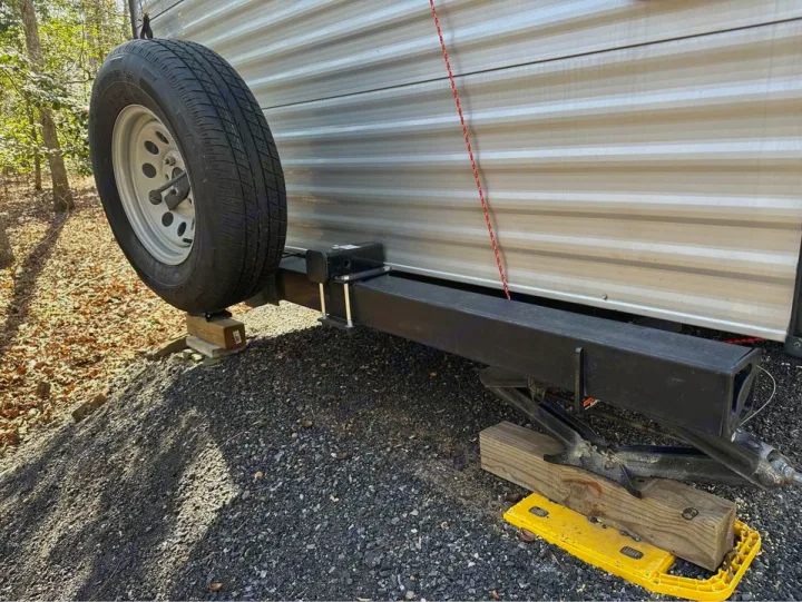 RV rental's rear: Spare tire on wood blocks. Jack and yellow leveling pads support the trailer on a gravel campsite.. Keystone RV Summerland Mini 2017