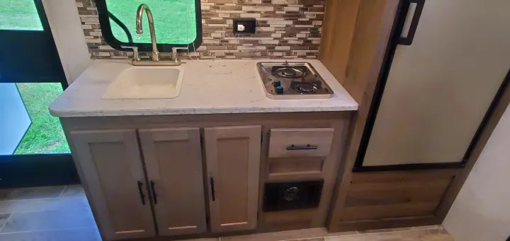 Compact RV rental kitchen features a sink, 2-burner stovetop, cabinets, and modern tiled backsplash.. Gulf Stream Trailmaster 2022