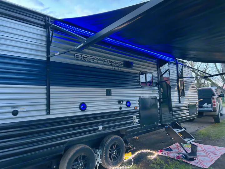 Experience an RV rental trip! This travel trailer features blue LED awning lights & an inviting outdoor setup at dusk.. Forest River Cherokee Grey Wolf 2022