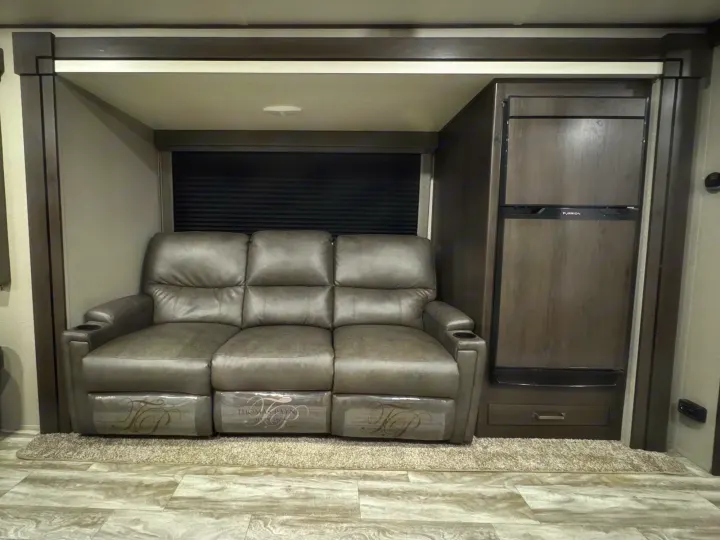 Cozy gray reclining sofa inside an RV rental, complete with a built-in refrigerator and wood accents.. Grand Design Transcend Xplor 2022