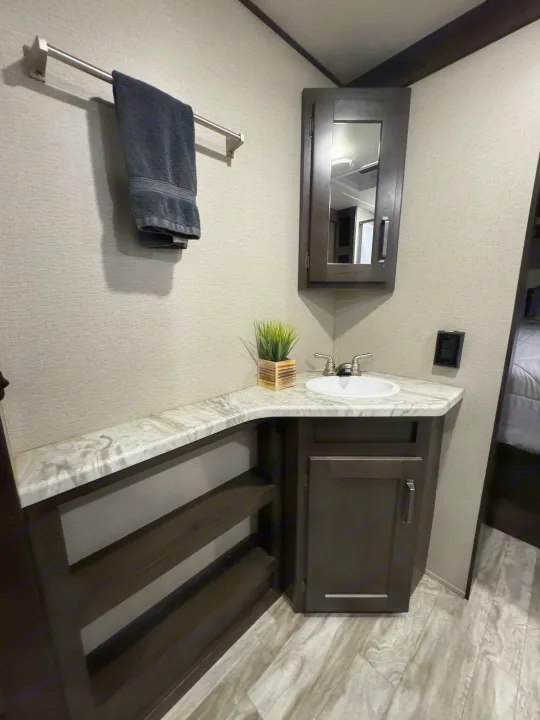Inside an RV rental: a compact bathroom vanity with white sink, marble-look counter, dark wood cabinets, towel bar, and plant.. Grand Design Transcend Xplor 2022